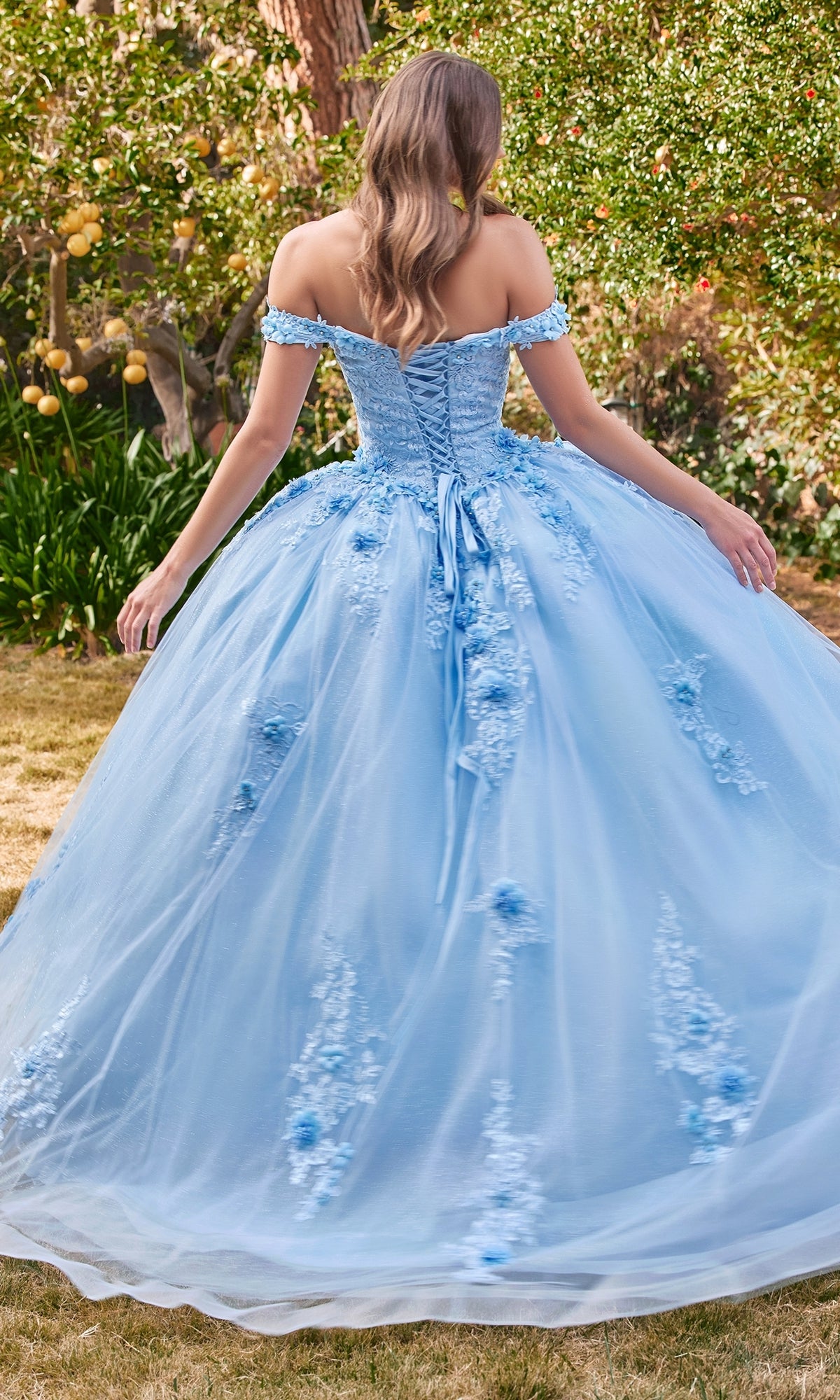 A woman with long hair stands outdoors, facing away, wearing the Ladivine 15702 Quinceañera Dress featuring light blue fabric, 3-D flowers, off-the-shoulder design, floral embroidery, and a lace-up back.